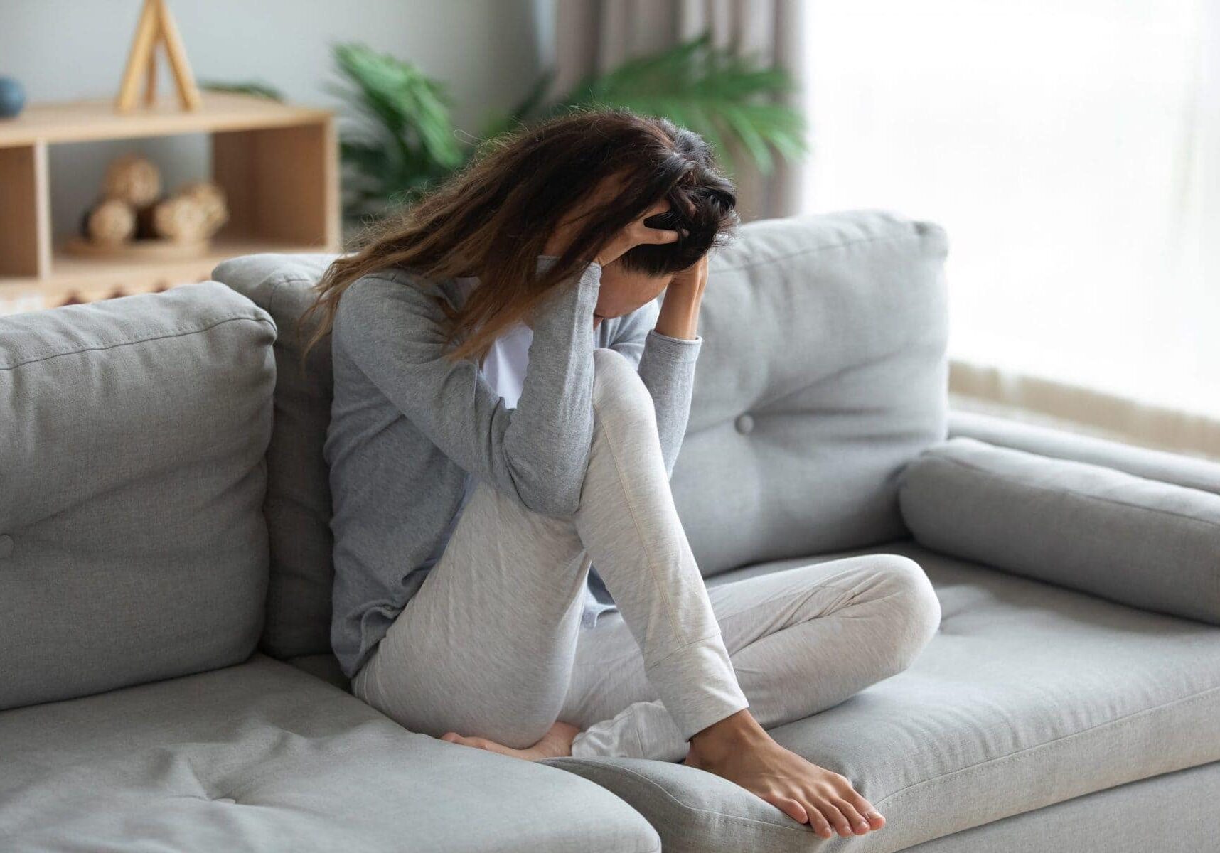 Stressful young mixed race woman sitting alone on sofa, holding head in hands, feeling desperate, suffering from relations break up or domestic violence. Unhealthy lady having strong headache.